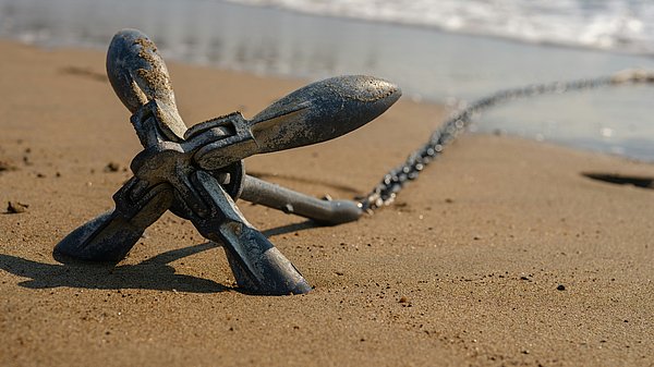 Metallanker mit Kette im Sand am Strand, im Hintergrund das Meer.