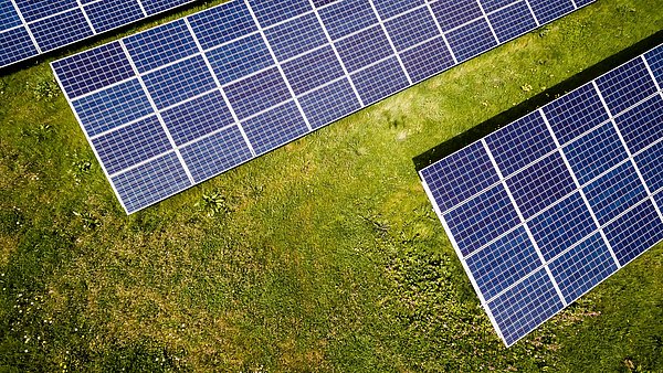 Mehrere Reihen von Solarmodulen stehen auf einer grünen Wiese. Die Aufnahme von oben zeigt die Anordnung der Photovoltaikmodule und die Vegetation zwischen den Anlagen.