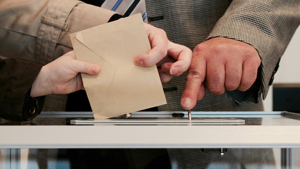 Mehrere Hände werfen Wahlumschläge in eine Wahlurne ein