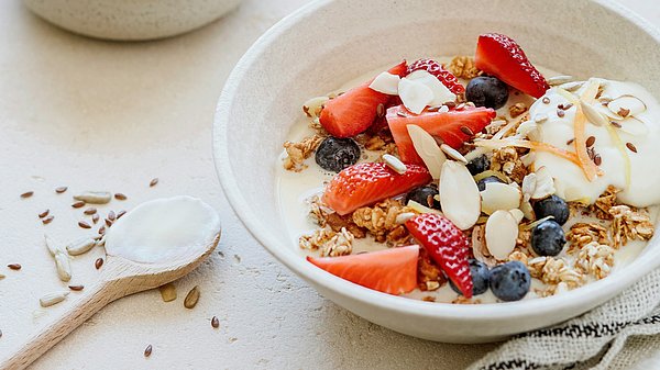 Müslischüssel mit Beeren und Nüssen, daneben ein Löffel und ein locker drapiertes Geschirrtuch auf dem Tisch.