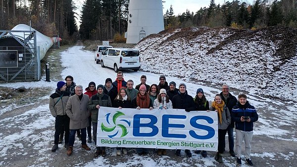 Gruppe von rund 20 Personen auf einem verschneiten Waldweg mit einem Banner „BES – Bildung Erneuerbare Energien Stuttgart e.V.“, im Hintergrund Windkraftanlage, Fahrzeuge und technische Anlagen.