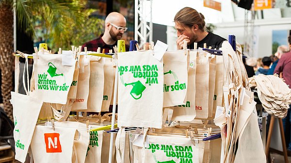 Siebdruckwerkstatt auf einer Messe: Jutetüten mit dem Aufdruck "Wir ernten was wir säen" sind aufgehängt zum Trocknen 