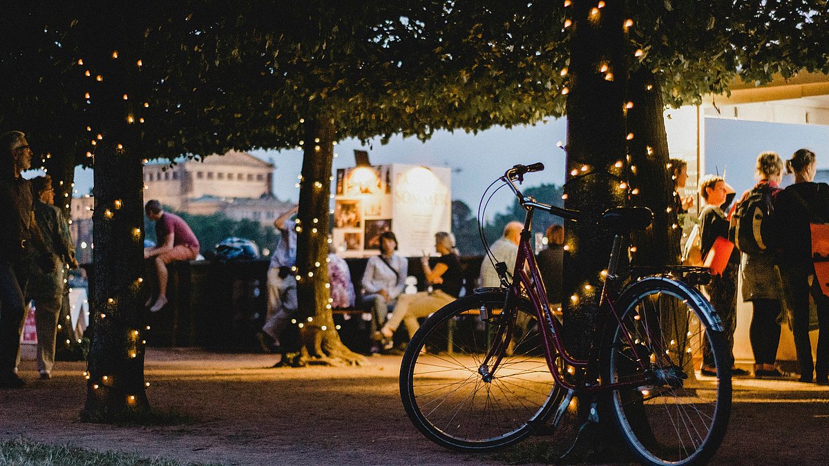 Fahrrad unter beleuchteten Bäumen bei einer Abendveranstaltung, Menschen im Hintergrund.