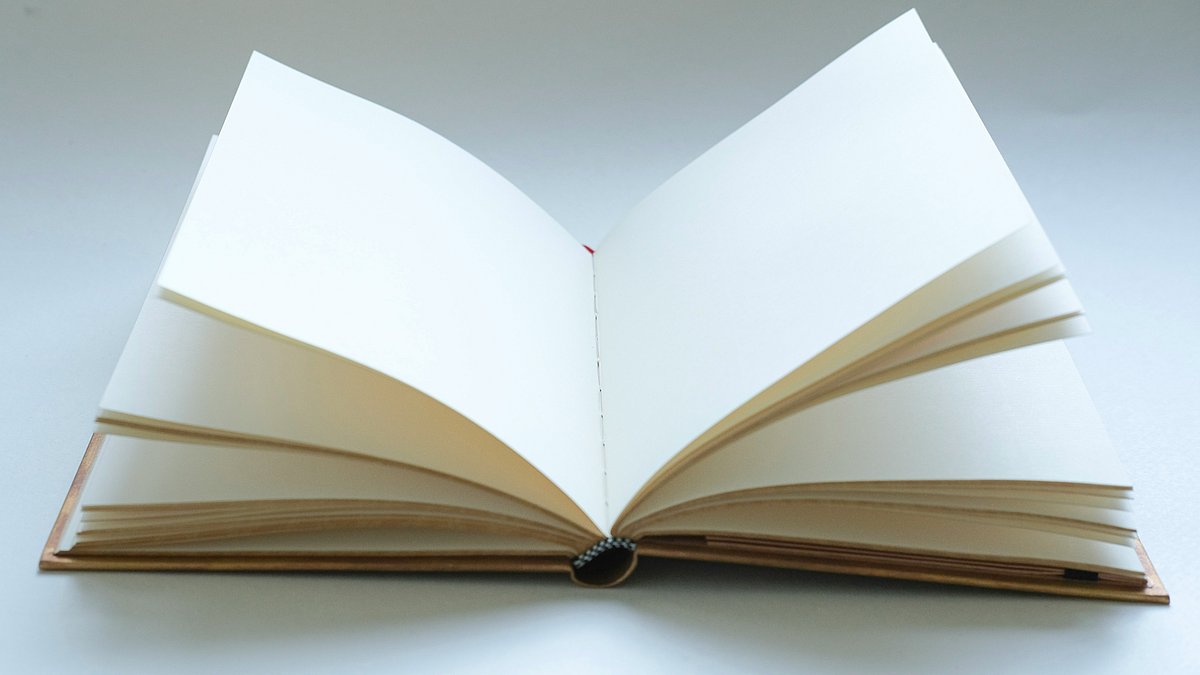 Aufgeschlagenes Buch mit leeren Seiten, das auf einer hellen Oberfläche liegt. Die sichtbare Fadenbindung in der Mitte und der schlichte Bucheinband veranschaulichen die handwerkliche Gestaltung eines gebundenen Buches.