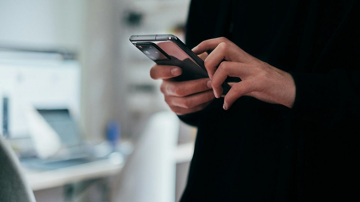 Nahaufnahme von Händen, die ein Smartphone bedienen, im Hintergrund ein unscharfer Schreibtisch.