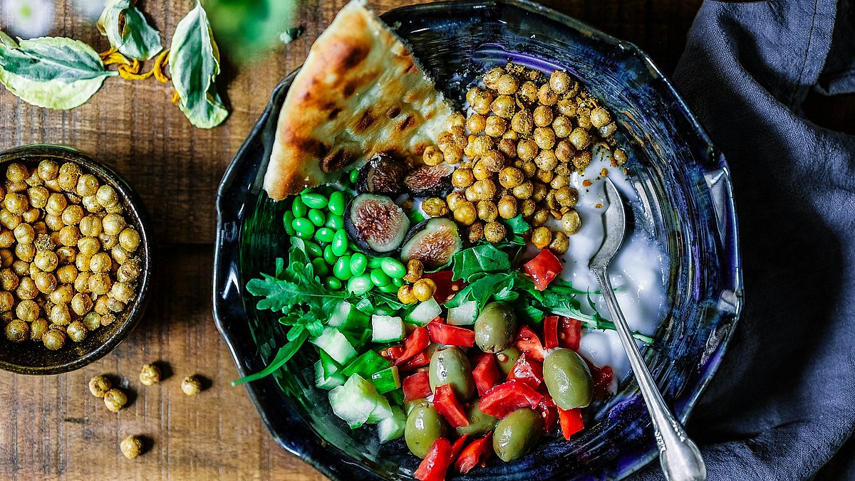 Schüssel mit buntem Salat aus Kichererbsen, Gemüse, Oliven und Fladenbrot auf einem Holztisch.