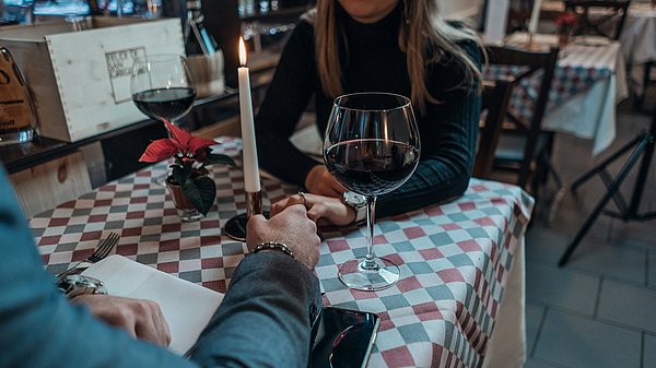 Zwei Personen halten über einen Tisch Händchen und trinken Rotwein