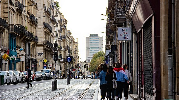 Menschen gehen gemeinsam durch eine belebte Innenstadtstraße mit Straßenbahnschienen, Geschäften und Wohnhäusern.