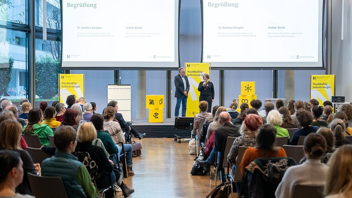 Teilnehmende sitzen in einem großen Raum vor einer Bühne, auf der Präsentationen gezeigt werden.