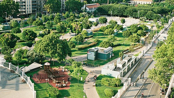 Großzügige Grünanlage in einem urbanen Umfeld mit zahlreichen Bäumen, Rasenflächen, Wegen und einem Spielplatz.