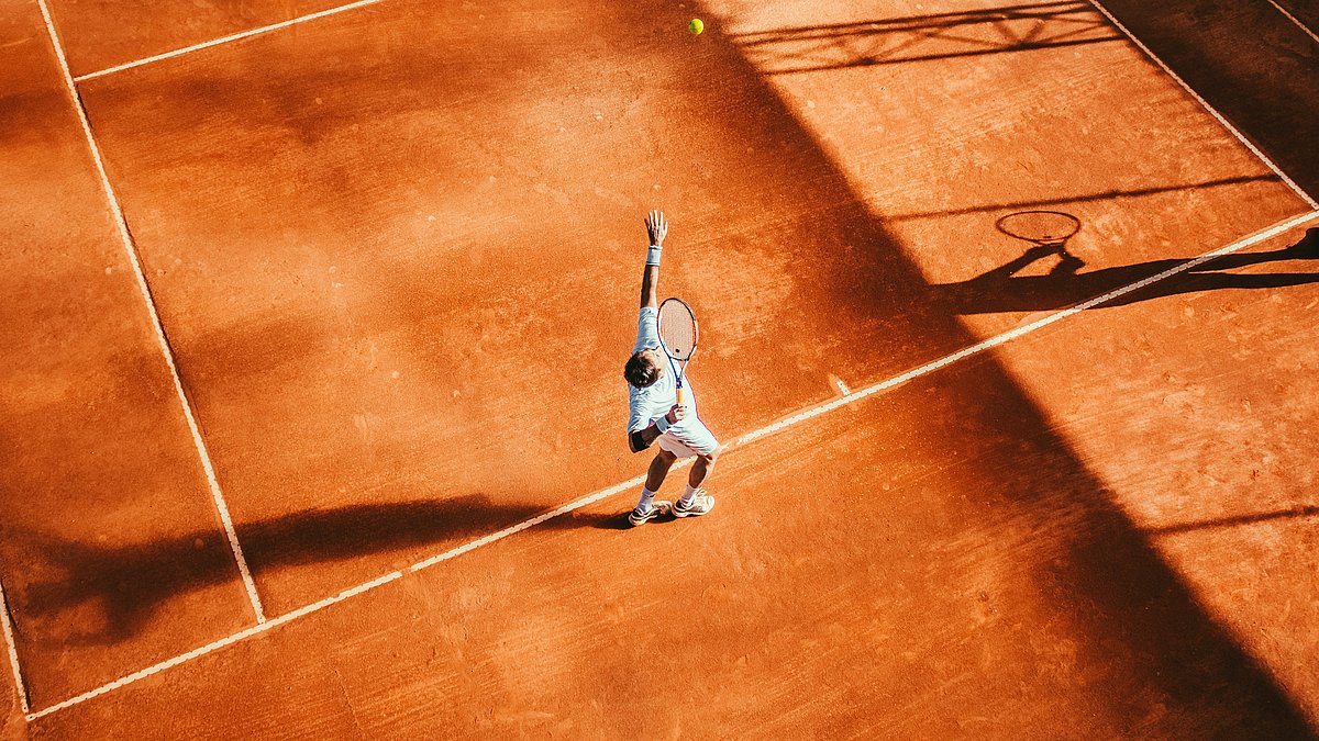 Person beim Aufschlag auf einem Tennis-Sandplatz, Ball in der Luft, lange Schatten.