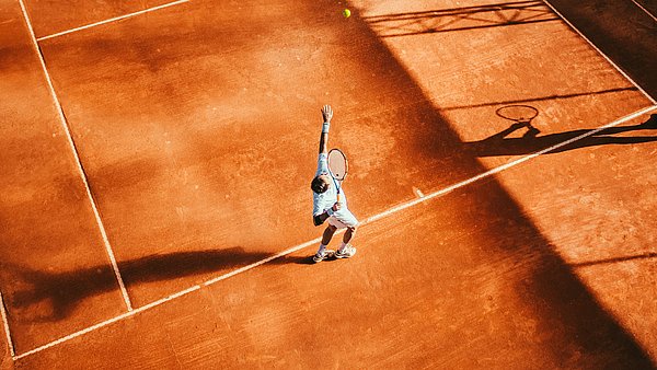 Person beim Aufschlag auf einem Tennis-Sandplatz, Ball in der Luft, lange Schatten.