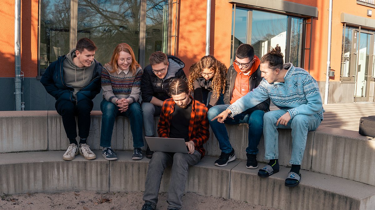 Mehrere junge Menschen sitzen gemeinsam auf einer Treppenstufe im Freien und betrachten einen Laptop.
