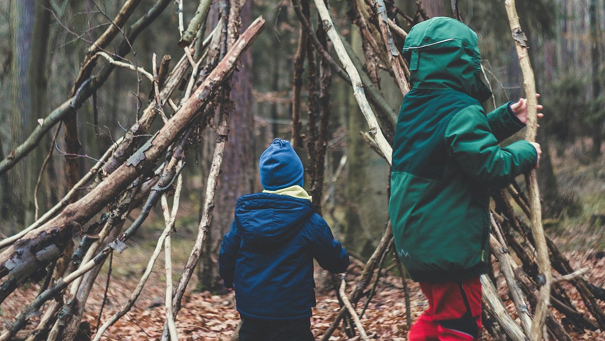 Kinder bauen aus Ästen und Zweigen eine einfache Hütte im Wald. Umgeben von Bäumen und Waldboden entsteht durch gemeinsames Arbeiten ein naturnaher Unterschlupf aus Naturmaterialien.