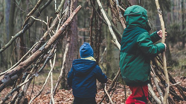 Kinder bauen aus Ästen und Zweigen eine einfache Hütte im Wald. Umgeben von Bäumen und Waldboden entsteht durch gemeinsames Arbeiten ein naturnaher Unterschlupf aus Naturmaterialien.